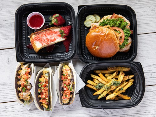 A variety of food items including tacos, a sandwich, fries, and dessert, arranged in black takeout containers.