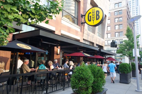 A bustling restaurant exterior with outdoor seating, umbrellas, and a prominent yellow sign, surrounded by city buildings.