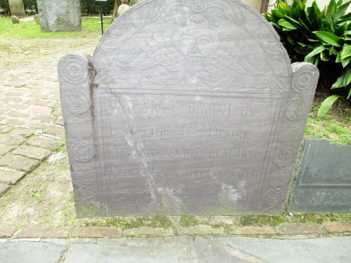 A weathered gravestone with engraved text, partially obscured, in a cemetery setting surrounded by greenery.