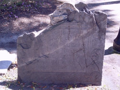 Weathered gravestone with faded inscriptions, partially broken, set in a park-like area with scattered leaves.