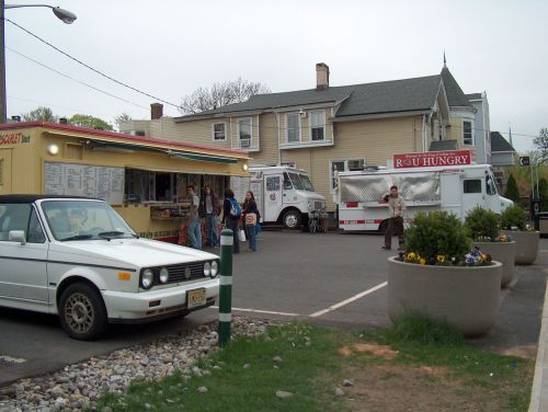 A food truck area with several trucks, a parked car, and people walking near a building on a cloudy day.