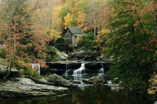 A rustic wooden mill beside a serene waterfall, surrounded by vibrant autumn foliage and rocky terrain.