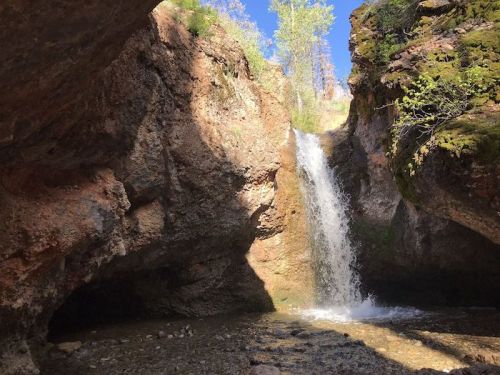 A serene waterfall cascades between rocky cliffs, surrounded by lush greenery under a clear blue sky.