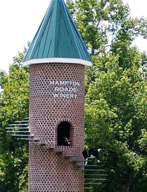 A tall tower with a green roof, labeled "Hampton Roads Winery," surrounded by trees and featuring a cow on a ledge.