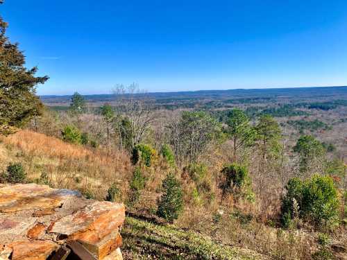 A scenic view of rolling hills and trees under a clear blue sky, with a stone wall in the foreground.