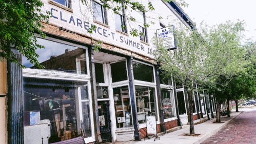 Historic storefront with large windows, trees lining the street, and a sign for Clarence T. Sumner, a local business.