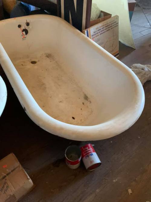 An old, dirty bathtub sits on a wooden floor, surrounded by clutter and a can of paint.