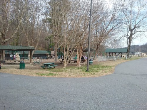 A playground with picnic areas and trees, set in a park with a paved path and a few people enjoying the space.