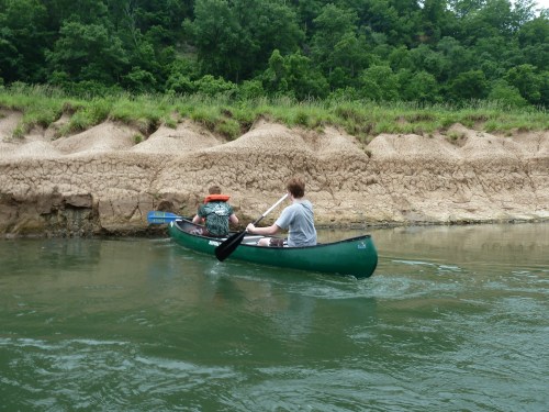 Two people paddle a green canoe along a river, with a sandy bank and lush greenery in the background.