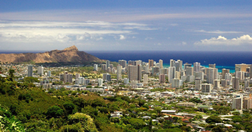 A panoramic view of a coastal city with skyscrapers, lush greenery, and a mountain in the background under a blue sky.