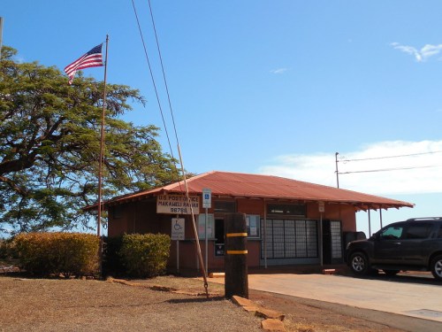 A small post office building with a red roof, U.S. flag, and a tree nearby under a clear blue sky.