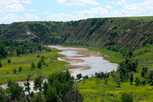 A winding river flows through lush green hills and cliffs under a bright blue sky with scattered clouds.