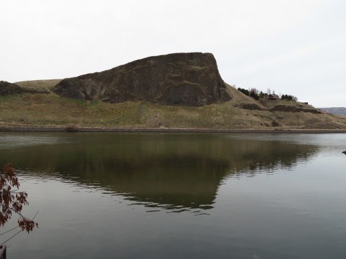 A calm lake reflects a rocky hill and grassy slopes under a cloudy sky.