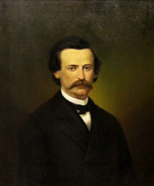 Portrait of a man with a mustache, wearing a black suit and bow tie, set against a dark, softly lit background.