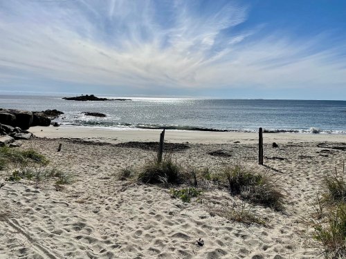 A serene beach scene with soft sand, gentle waves, and a clear blue sky, featuring distant rocks on the horizon.