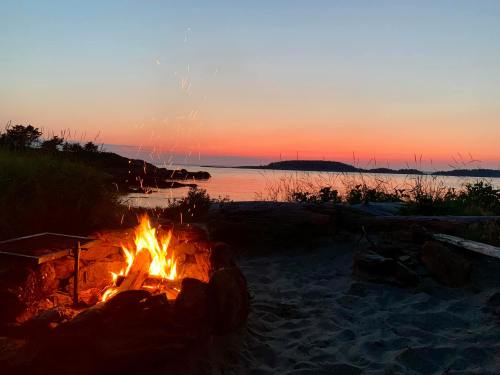A cozy campfire glows against a serene sunset over the water, with silhouettes of distant islands and grassy shorelines.
