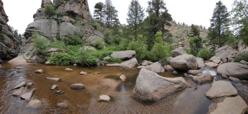 A serene landscape featuring a rocky riverbank surrounded by lush greenery and towering cliffs under a cloudy sky.