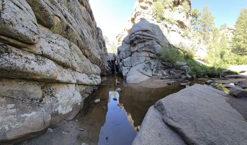 A narrow canyon with rocky walls and a calm pool of water reflecting the surrounding greenery and blue sky.