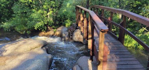 A wooden bridge over a flowing stream, surrounded by lush greenery and large rocks.
