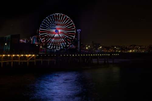 A brightly lit Ferris wheel on a pier at night, reflecting in the water under a dark sky.