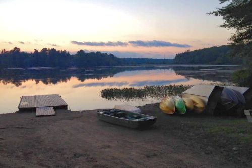 Serene lakeside scene at sunset with a dock, a small boat, and colorful kayaks on the shore.