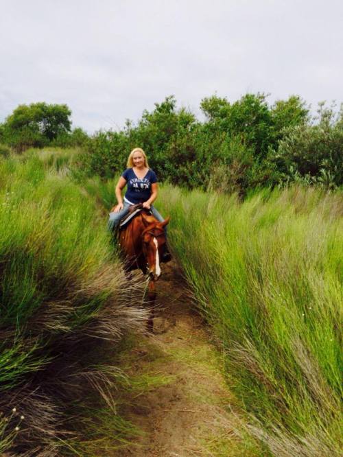 A woman riding a brown horse along a narrow path surrounded by tall green grass and shrubs.