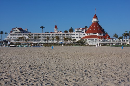 Historic hotel with a red roof, palm trees, and sandy beach under a clear blue sky.