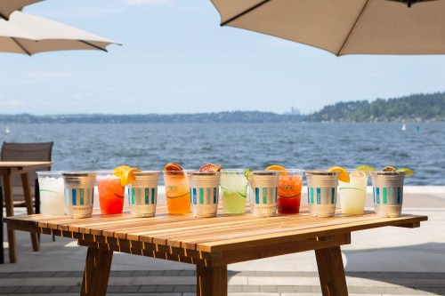 A wooden table by the water displays a variety of colorful drinks under large umbrellas.