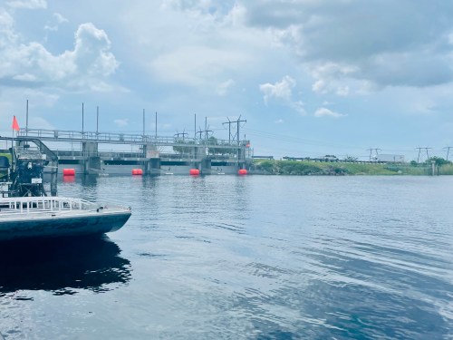 A boat in the foreground with a dam and power lines in the background under a partly cloudy sky.