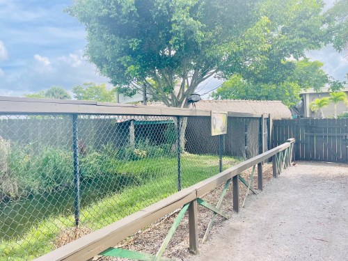 A pathway with a wooden railing beside a fenced area, featuring greenery and a tree under a cloudy sky.