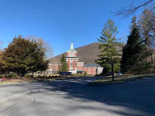 A scenic view of a large brick building surrounded by trees and mountains under a clear blue sky.