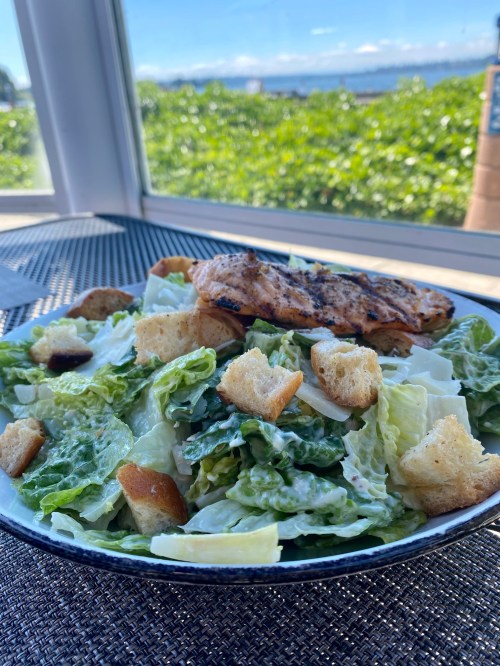 A fresh Caesar salad with grilled chicken and croutons, served on a plate with a scenic outdoor view.