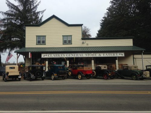 A vintage general store with several classic cars parked in front, set against a cloudy sky.