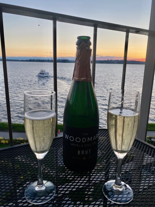 A bottle of sparkling wine with two glasses on a balcony overlooking a sunset over the water.