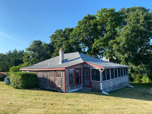 A cozy, single-story house with a screened porch, surrounded by green trees and a grassy lawn under a clear blue sky.