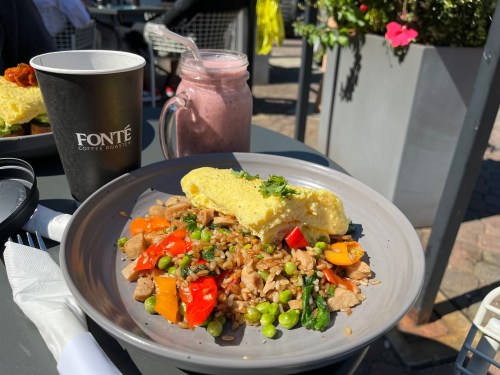 A plate with an omelette on a bed of colorful fried rice, alongside a smoothie and a coffee cup on a sunny outdoor table.