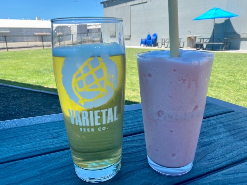 A glass of beer and a pink smoothie sit on a table outdoors, with green grass and a blue umbrella in the background.