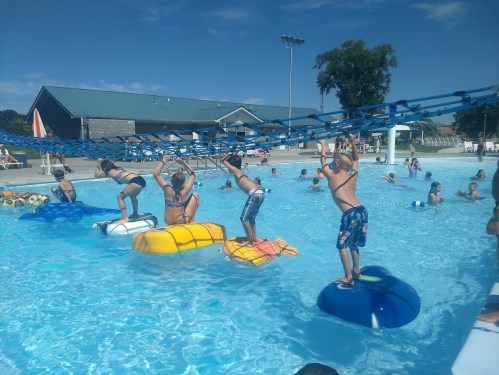 Children play on colorful floats in a pool, navigating a rope obstacle above the water on a sunny day.