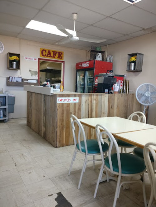 A cozy café interior with a wooden counter, tables, and a soda machine, featuring a fan and decorative plants.