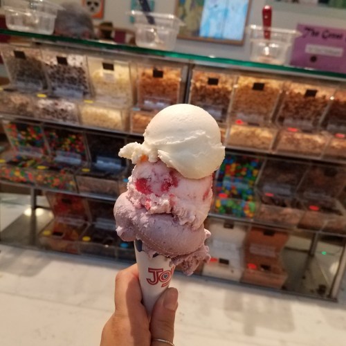 A hand holding a colorful ice cream cone with three scoops in front of a display of various toppings.