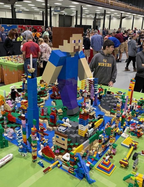 A large LEGO figure stands over a colorful LEGO cityscape, with people browsing in the background at a crowded event.
