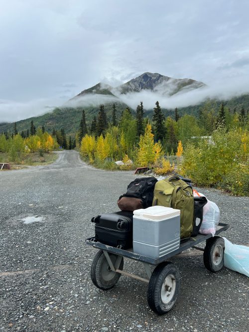 A cart loaded with gear sits on a gravel path, surrounded by trees and mountains shrouded in mist.