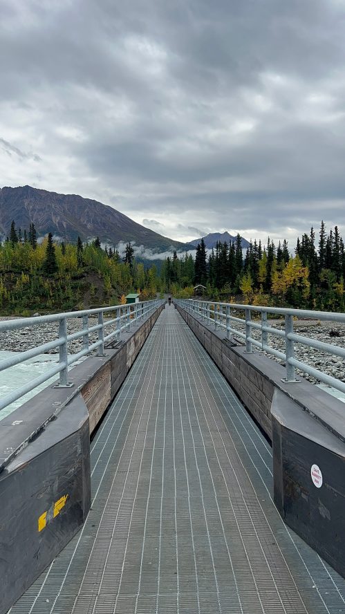 A long walkway stretches through a forested area, with mountains in the background under a cloudy sky.