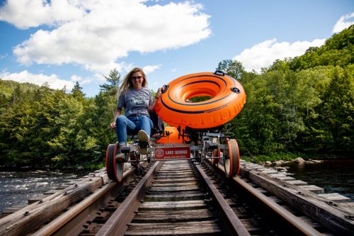A woman sits on a rail cart with an orange inflatable tube, surrounded by trees and a river under a blue sky.