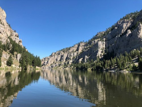 A serene river surrounded by tall cliffs and lush trees, reflecting the clear blue sky and rocky landscape.
