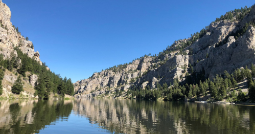 A serene river surrounded by steep cliffs and lush greenery under a clear blue sky. Reflections shimmer on the water.