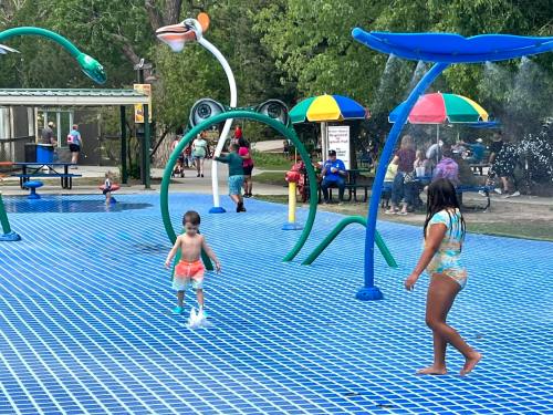 Children play in a colorful splash pad, with water features and umbrellas in a park setting.