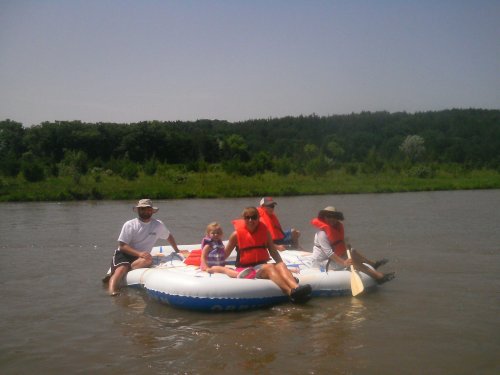 A group of four people and a child in life jackets relax on an inflatable raft on a river surrounded by greenery.