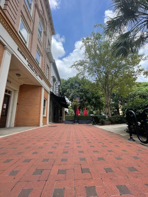 A brick pathway leads to a clock tower surrounded by trees and outdoor seating with red umbrellas under a blue sky.