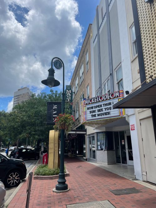 A street view of a movie theater with a marquee, surrounded by buildings and trees under a partly cloudy sky.